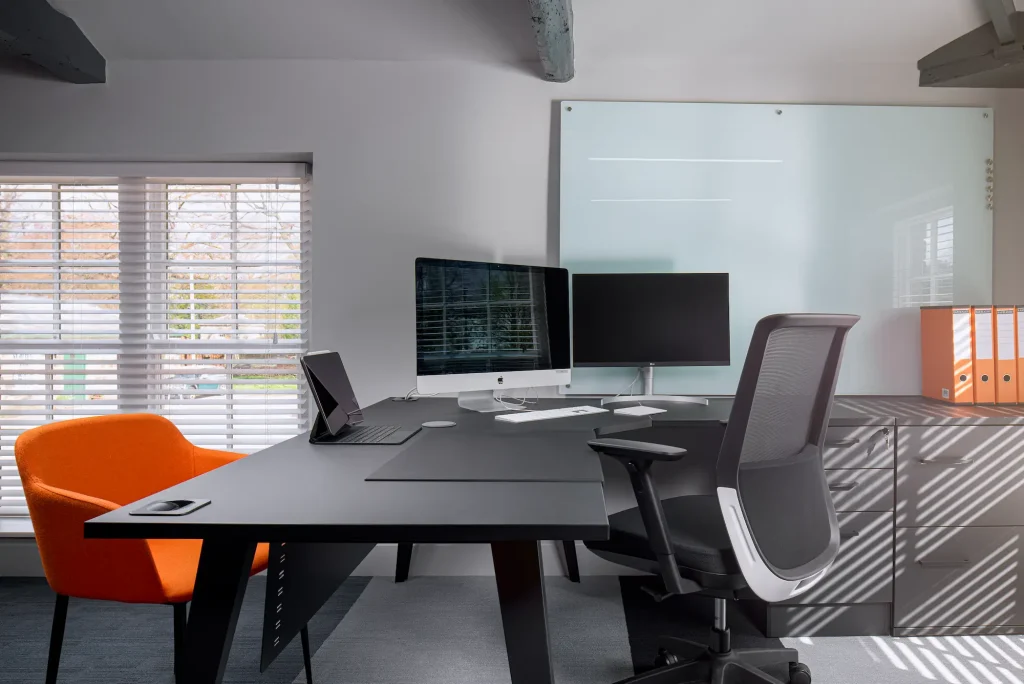 Modern desk space with grey decor, with an orange accent chair to the left of the desk. The room is well-lit with natural light and has minimalistic, professional design elements and a large white board on the wall of the office space.